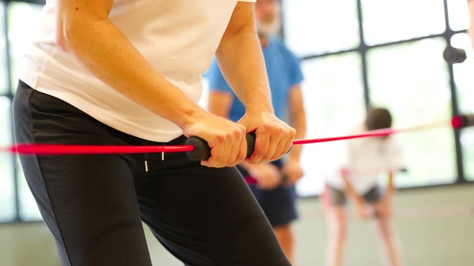 Nahaufnahme einer Person mit einem Widerstandsband im Fitnessstudio, umgeben von weiteren trainierenden Personen.