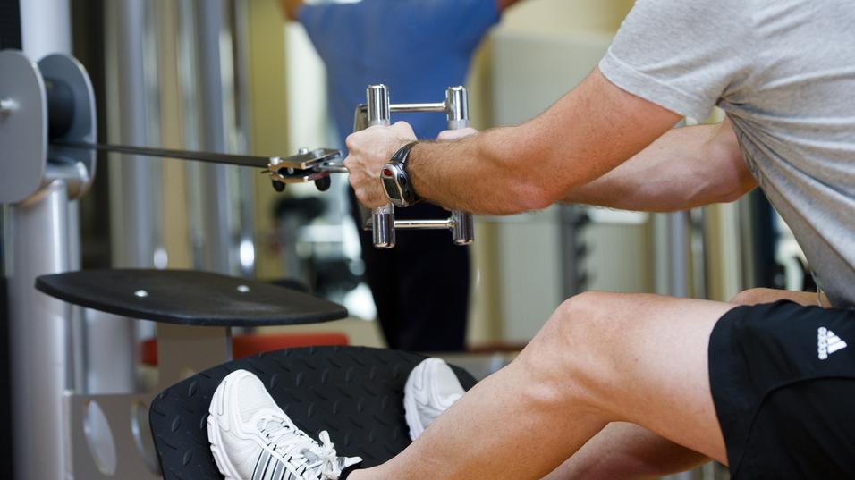 Mann trainiert im Fitnessstudio mit einem Kabelzuggerät, ein anderer Mensch im Hintergrund führt eine Übung aus.