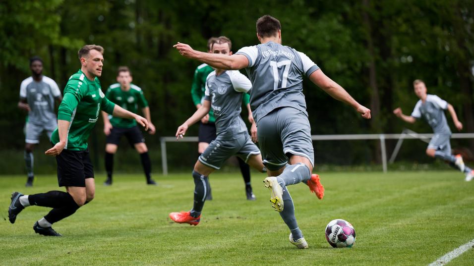 Drei Fußballspieler in grünen und grauen Trikots auf einem Rasenplatz, während ein Spieler den Ball zuspielt.
