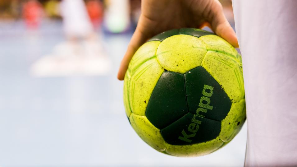 Nahaufnahme einer Hand, die einen grünen Handball mit gelben Elementen hält, im Hintergrund Spieler auf dem Spielfeld.