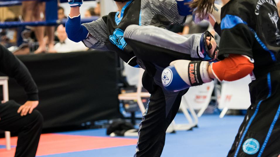 Zwei Kämpferinnen im Kickboxen, eine tritt mit dem Bein aus einem Kampfstand auf einen Mattenboden.