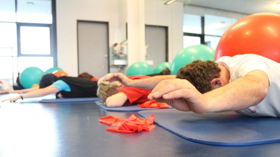 Gruppentraining mit Personen liegend auf Matten, während Bälle im Hintergrund sichtbar sind.