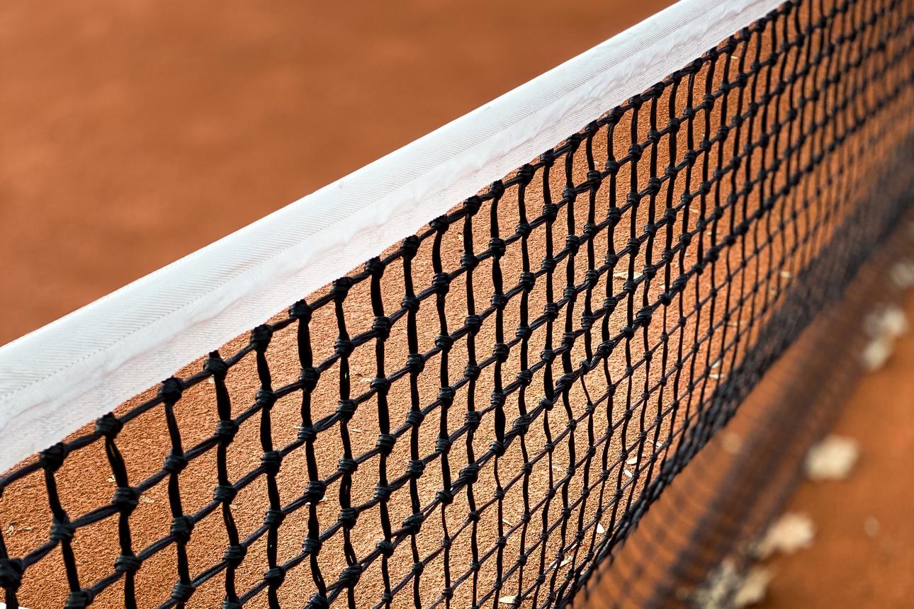 Nahaufnahme eines Tennisnetzes auf einem roten Sandplatz mit Blättern im Hintergrund.