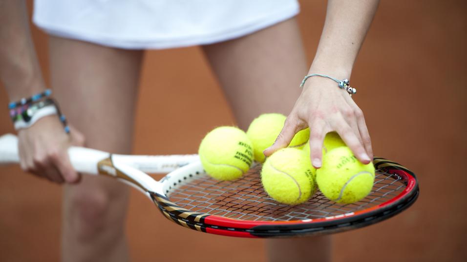 Hand greift mehrere gelbe Tennisbälle auf einem Tennisschläger, während die andere Hand die Schlägerhaltung stabilisiert.